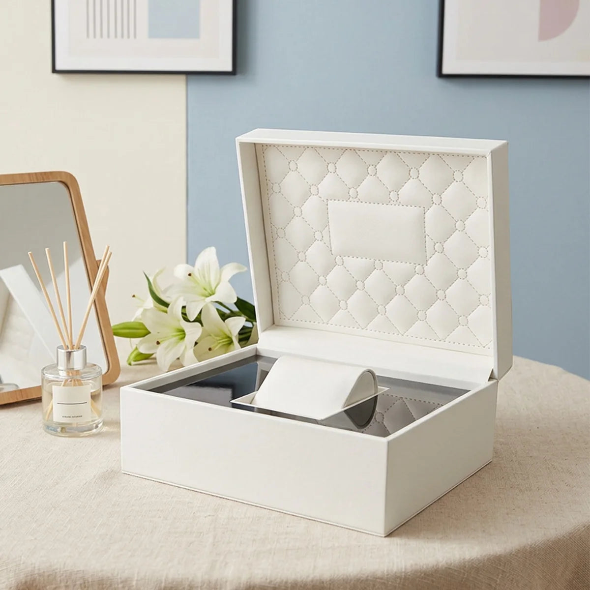 Boîte à montre Le Matelassé, coque ivoire et intérieur matelassé sur une table à côté d'un diffuseur de parfum et d'un miroir